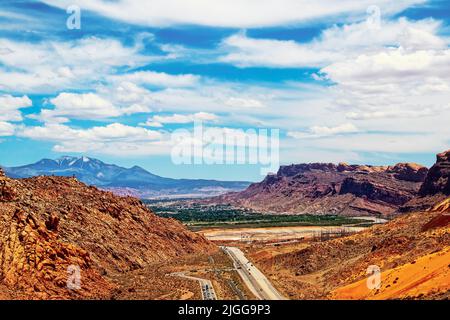 Blick auf Moab, Utah, und die Berge dahinter, und Autos stehen auf dem Gipfel des Arches National Park an, um den Park zu betreten Stockfoto