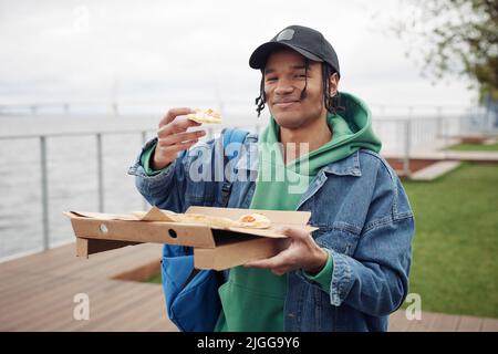 Fröhlicher schwarzer Kerl in der Freizeitkleidung, der ein Stück Pizza über einer quadratischen Schachtel hält, während er einen Snack hat und während des Picknicks im Park die Kamera anschaut Stockfoto