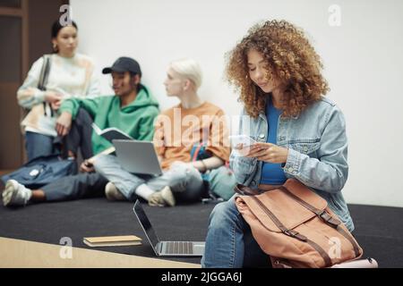 Hübsches Teenager-Mädchen in Casualwear, das in der Pause in einem Smartphone schreibt, während sie im Hörsaal sitzt, während ihre Gruppenmitglieder über Vortragsnotizen diskutieren Stockfoto