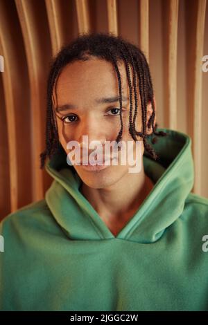 Nahaufnahme eines ruhigen afroamerikanischen Teenagers im grünen Hoodie, der die Kamera anschaut, während er gegen die beige Wand steht Stockfoto