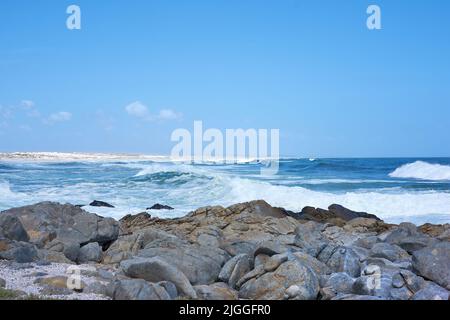 Felsen im Meer unter einem blauen Himmel mit Kopierraum. Schöne Landschaft von Strandwellen, die gegen Felsbrocken oder große Steine im Meer bei A plantschen Stockfoto