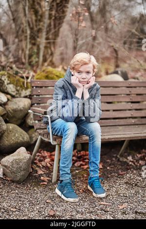 Gelangweilter Teenager, der alleine draußen auf einer Parkbank sitzt und sich traurig und einsam fühlt. Ein junger ernsthafter männlicher Junge oder ein Kind draußen mit den Händen auf dem Kinn Stockfoto