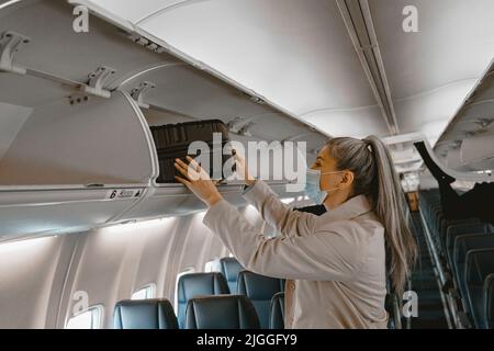 Frau in Maske, die Gepäck während des Boarding in den Gepäckraum im Flugzeug legt Stockfoto