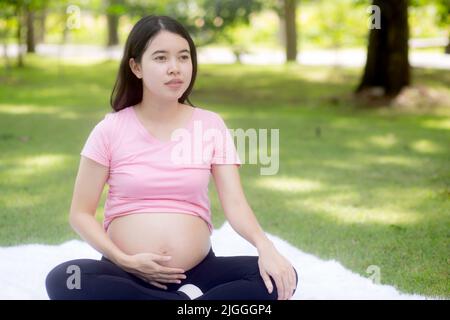 Glück junge asiatische Frau schwanger sitzen Yoga mit Meditation Lotus zur Entspannung im Park, Mutter Training für die Gesundheit in der üben Stockfoto