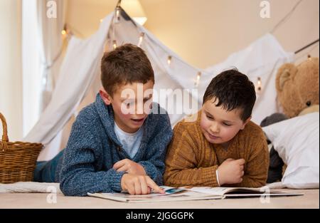 Diese kleinen Jungen genießen die gemeinsame Geschichte. Zwei Brüder lesen zu Hause unter einer Decke ein Buch. Stockfoto