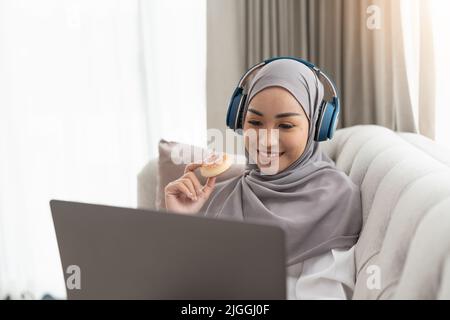 Eine schöne, entspannte asiatische muslimische Frau, die einen Hijab und Kopfhörer trägt, schaut sich einen Film auf ihrem Laptop-Computer an, während sie im Wohnzimmer einen heißen Tee trinkt Stockfoto