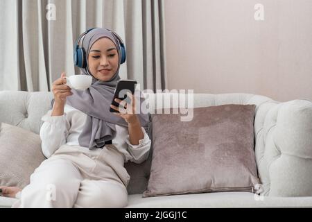 Asiatischer muslim, der Kopfhörer trägt und Musik vom Mobiltelefon auf dem Sofa zu Hause hört. Stockfoto