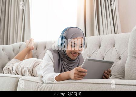 Ganzkörperansicht einer jungen muslimischen Frau, die im Wohnzimmer auf der Couch liegt und sich einen Film auf einem digitalen Tablet ansieht Stockfoto