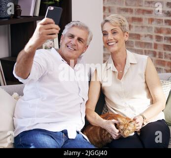 Ein reifes Paar, das Selfies macht, während es sich mit seiner Katze zu Hause auf einem Sofa entspannt. Stockfoto