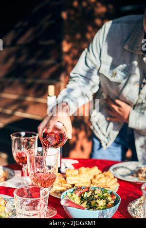 Eine Frau gießt Wein in Gläser. Ein voller Tisch ist für ein freundliches Bankett im Freien aufgestellt. Vertikales Foto. Stockfoto