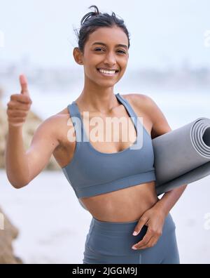 Ich bin auch unten für eine gute Yoga-Sitzung. Ein beschnittenes Porträt einer attraktiven jungen Frau, die beim Yoga am Strand Daumen nach oben gibt. Stockfoto