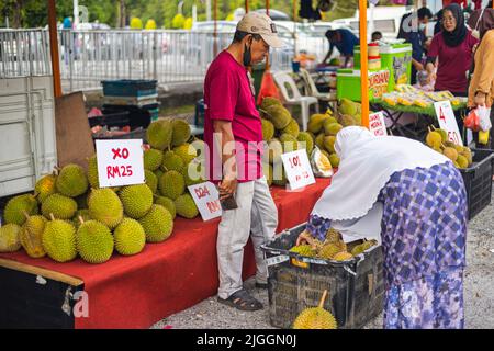 Kuala Lumpur, Malaysia - 21. Juni 2022: Ein elderlicher Mann auf einem Straßenmarkt verkauft Durian. Eine muslimische Frau mit Hijab, die in einem Korb nach dem richtigen f sucht Stockfoto