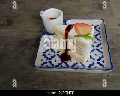 Erdbeer- und Kiwi-Fruchtscheibe mit rotem Sirup und cremefarbenem Biskuitkuchen in einer Keramikschale auf einem grauen Holztisch, heißer Espresso-Kaffee in weißer Tasse Stockfoto