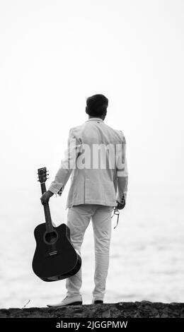 Weiß gekleideter Mann, der eine Gitarre in der Hand hält, Blick von hinten in den Strand, Schwarz-Weiß-Fotografie. Stockfoto