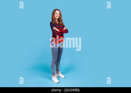 In voller Länge Porträt einer gut aussehenden Frau mit gestreiftem Pullover im lässigen Stil und Jeans, die mit gekreuzten Armen stehen und die Kamera betrachten. Innenaufnahme des Studios isoliert auf blauem Hintergrund. Stockfoto