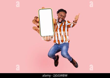 Wie und fliegen. Glücklicher junger lustiger Mann Daumen nach oben, fliegen und springen in der Luft und zeigen große mobile leere Bildschirm für Kopieplatz und Werbebereich. Indoor-Studio auf rosa Hintergrund gedreht Stockfoto