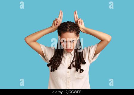 Porträt einer tyrannisierenden Frau mit schwarzen Dreadlocks, die bedrohlich aussehen und ein Bullenhornschild zeigen, die Finger auf dem Kopf halten, eine widerstreitende Person. Innenaufnahme des Studios isoliert auf blauem Hintergrund. Stockfoto