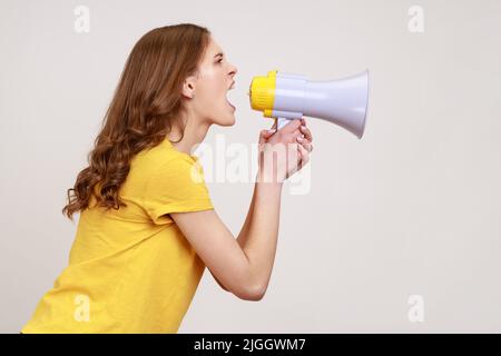 Profil Porträt nervös braunhaarige Frau jungen Alters in gelben T-Shirt laut sprechend schreiend hält Megaphon, kündigt wichtige Botschaft. Innenaufnahme des Studios isoliert auf grauem Hintergrund. Stockfoto