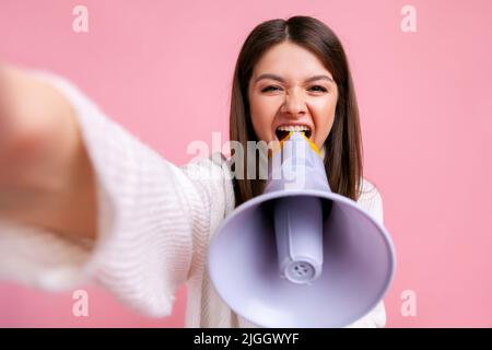 Aufgeregt Brünette weibliche Schreien laut in Megaphon, so dass Standpunkt Foto aus der Präsentation, trägt weißen lässigen Stil Pullover. Innenaufnahme des Studios isoliert auf rosa Hintergrund. Stockfoto