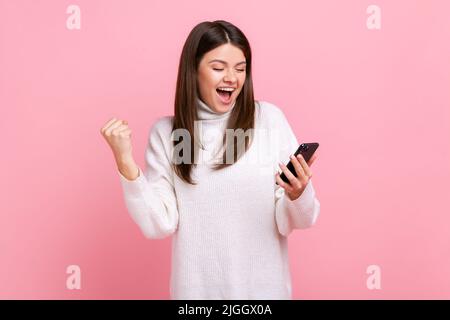 Happy positive Mädchen hält Smartphone in den Händen, kleckende Faust, feiert Sieg, plötzlichen Erfolg, trägt weißen lässigen Stil Pullover. Innenaufnahme des Studios isoliert auf rosa Hintergrund. Stockfoto