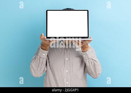 Unbekannter Mann versteckt sich hinter dem Laptop mit weißem, leerem Display und präsentiert neue Anwendungen, präsentiert Werbefläche, trägt gestreiftes Hemd. Innenaufnahme des Studios isoliert auf blauem Hintergrund. Stockfoto