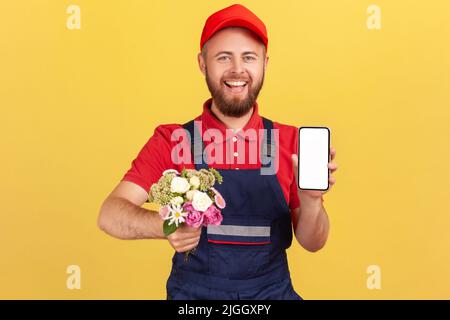 Lächelnder, positiver Mann, Kurier, der mit Blumen in den Händen stand und das Handy mit leerem Bildschirm für Werbung zeigte und auf die Kamera schaute. Innenaufnahme des Studios isoliert auf gelbem Hintergrund. Stockfoto