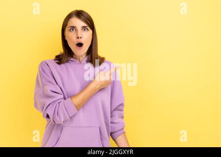 Porträt einer aufgeregten, schönen jungen erwachsenen Frau, die mit einem überraschten Gesicht auf den Copyspace zeigt und einen violetten Hoodie trägt. Innenaufnahme des Studios isoliert auf gelbem Hintergrund. Stockfoto