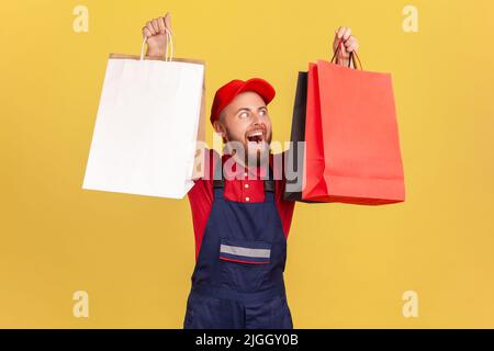 Porträt eines zufriedenen bärtigen Lieferers in blauer Uniform, der Papier-Einkaufstaschen hält, zufrieden mit dem großen Verkauf am schwarzen freitag, der Ordnung bringt. Innenaufnahme des Studios isoliert auf gelbem Hintergrund. Stockfoto