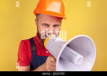 Wütender, aggressiver bärtiger Arbeiter mit Schutzhelm und blauen Overalls, der Megaphon hält und schreiendes, aus der Sicht gehaltenes Foto. Innenaufnahme des Studios isoliert auf gelbem Hintergrund. Stockfoto