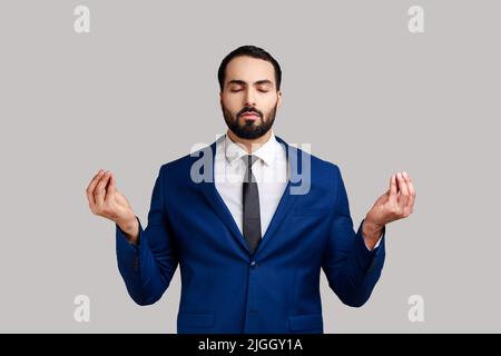 Porträt eines entspannten, gutaussehenden bärtigen Mannes, der mit erhobenen Armen steht und Yoga-Meditationsübungen macht und einen Anzug im offiziellen Stil trägt. Innenaufnahme des Studios isoliert auf grauem Hintergrund. Stockfoto