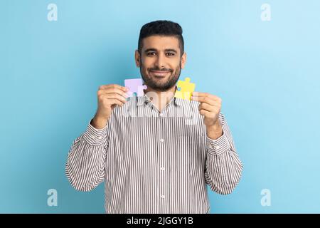 Porträt eines optimistischen bärtigen Geschäftsmannes, der gelbe und violette Puzzleteile hält, Aufgaben löst, die Kamera anschaut und ein gestreiftes Hemd trägt. Innenaufnahme des Studios isoliert auf blauem Hintergrund. Stockfoto