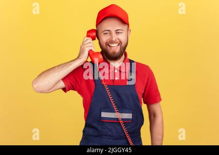 Porträt eines lächelnden freundlichen Arbeiters in blauen Overalls, der auf rotem Retro-Telefon mit Kunden spricht, Aufträge annimmt, professionelle Dienstleistungsbranche. Innenaufnahme des Studios isoliert auf gelbem Hintergrund. Stockfoto