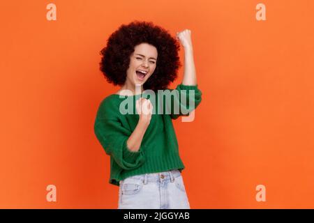 Porträt einer glücklichen, aufgeregten Frau mit Afro-Frisur im grünen Pullover im lässigen Stil, die ihre siegreichen, geballten Fäuste feiert und brüllt. Innenaufnahme des Studios isoliert auf orangefarbenem Hintergrund. Stockfoto