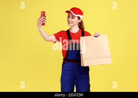 Porträt der Lieferung Frau posiert mit Papierpaket in den Händen, Mobiltelefon für die Kommunikation per Videoanruf, tragen Overalls und rote Kappe. Innenaufnahme des Studios isoliert auf gelbem Hintergrund. Stockfoto