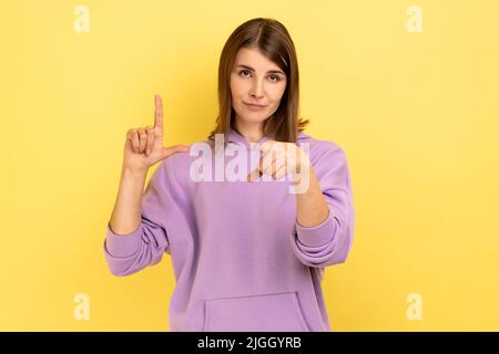 Porträt einer frustrierten jungen Frau, die eine lockere Geste zeigt und auf die Kamera zeigt, mit einem mürrischen Gesicht und einem violetten Hoodie blickt. Innenaufnahme des Studios isoliert auf gelbem Hintergrund. Stockfoto