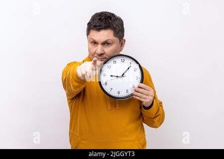 Porträt eines glücklichen, positiven Mannes, der eine große Wanduhr hält und mit einem angenehmen Lächeln auf die Kamera zeigt, Zeit zu gehen und einen Kapuzenpullover im urbanen Stil trägt. Innenaufnahme des Studios isoliert auf weißem Hintergrund. Stockfoto