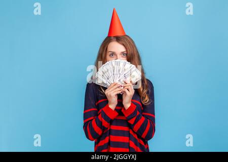 Schüchterne Frau mit gestreiftem Pullover im lässigen Stil und rotem Partykegel, die sich hinter Geldscheinen versteckt, genießt das reiche Leben und ist mit dem Geldgeschenk zufrieden. Innenaufnahme des Studios isoliert auf blauem Hintergrund. Stockfoto
