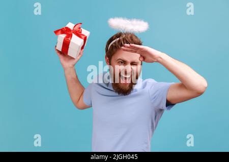 Porträt eines positiv optimistischen bärtigen Engelsmenschen mit einem heiligen Nimbus, der eine verpackte Geschenkbox hält und weit schaut, zwinkert und wegschaut. Innenaufnahme des Studios isoliert auf blauem Hintergrund. Stockfoto