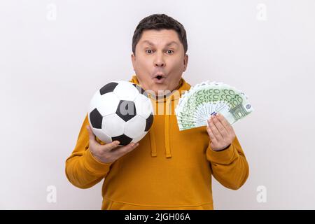 Schockierter Mann mittleren Alters, der Fußball zeigt und sich über hundert Euro-Scheine lustig macht, viel Geld für Sport gewinnt und einen Kapuzenpullover im urbanen Stil trägt. Innenaufnahme des Studios isoliert auf weißem Hintergrund. Stockfoto