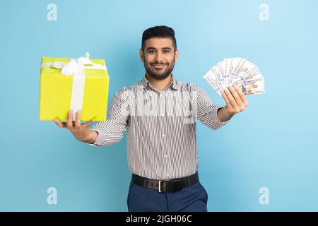 Porträt eines glücklichen Geschäftsmannes, der eine eingepackte Schachtel aushält, und eines großen Fans von Dollar-Banknoten, der die Kamera anschaut und ein gestreiftes Hemd trägt. Innenaufnahme des Studios isoliert auf blauem Hintergrund. Stockfoto