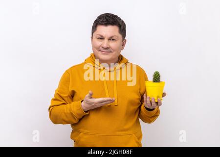 Porträt eines zufriedenen, positiven Mannes, der einen gelben Blumentopf und Kaktus präsentiert, mit einem toothy Lächeln auf die Kamera blickt und einen Kapuzenpullover im urbanen Stil trägt. Innenaufnahme des Studios isoliert auf weißem Hintergrund. Stockfoto
