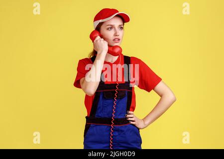 Staunende Arbeiterin, die auf einem roten Retro-Telefon mit Kunden spricht, wegschaut, professionelle Dienstleistungsbranche, Overalls und rote Kappe trägt. Innenaufnahme des Studios isoliert auf gelbem Hintergrund. Stockfoto