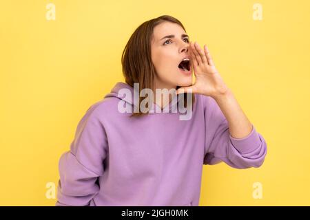 Achtung, Werbung. Profil einer Frau, die laut über den Verkauf schreit, eine wichtige Botschaft ankündigt und einen violetten Hoodie trägt. Innenaufnahme des Studios isoliert auf gelbem Hintergrund. Stockfoto