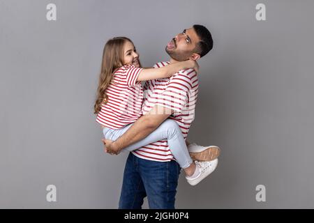 Porträt eines lustigen Vaters und einer lächelnden charmanten Tochter in gestreiften T-Shirts, die zusammen stehen, eines müden Vaters, der das Kind hält und die Zunge zeigt. Innenaufnahme des Studios isoliert auf grauem Hintergrund. Stockfoto