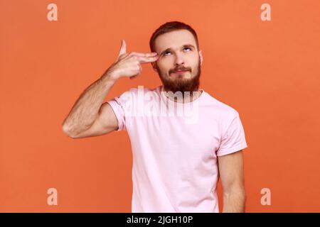 Selbstmordgesten. Porträt eines frustrierten, verzweifelten bärtigen Mannes, der mit der Fingerpistole auf den Kopf zeigt, sich deprimiert fühlt und sich mit einem T-Shirt schießt. Innenaufnahme des Studios isoliert auf orangefarbenem Hintergrund. Stockfoto