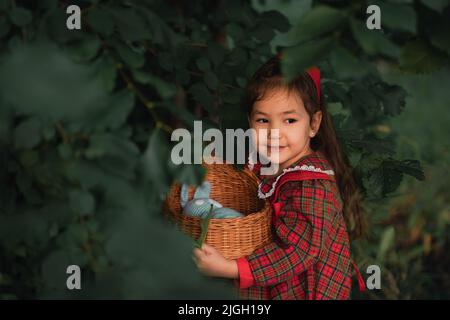 Kleines Mädchen in rotem Kleid hält Weidenwiege in den dicken Büschen Stockfoto