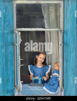 Porträt eines Schneidermädchen Nähen Sie Puppenkleidung auf einer Kinder-Nähmaschine im Fenster eines alten Holzhauses. Stockfoto