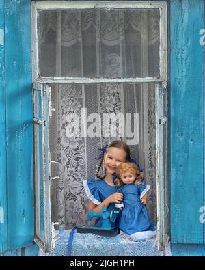 Porträt eines Schneidermädchen Nähen Sie Puppenkleidung auf einer Kinder-Nähmaschine im Fenster eines alten Holzhauses. Stockfoto