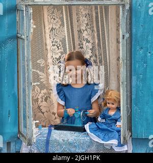 Porträt eines Schneidermädchen Nähen Sie Puppenkleidung auf einer Kinder-Nähmaschine im Fenster eines alten Holzhauses. Stockfoto