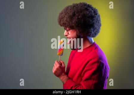 Seitenansicht eines Hipster-Mannes mit Afro-Frisur und Eis in den Händen, geniesst das Lecken eines leckeren Desserts und trägt ein rotes Sweatshirt. Innenaufnahmen im Studio, isoliert auf farbigem Neonlicht-Hintergrund. Stockfoto